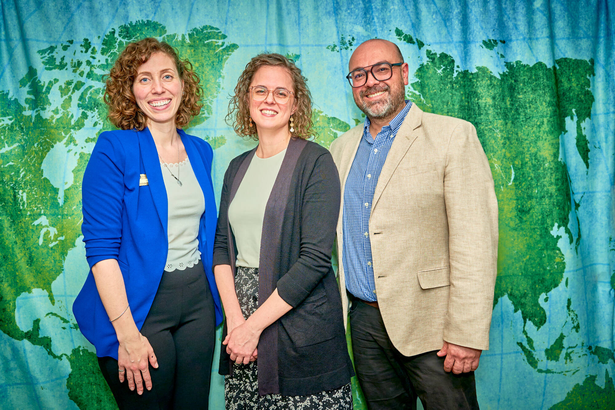 Three staff members of International Student Services standing smiling in front of a printed world backdrop